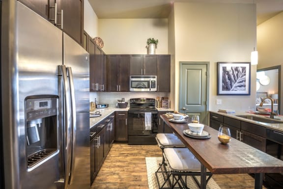 Kitchen with dark wood cabinets, stainless microwave, refrigerator, black stove, a walk-in pantry, and granite countertop. Island with two barstools and decorations and view into bedroom.