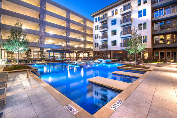 Courtyard pool area during nighttime with lights in water and view of surrounding apartments and balconies and parking garage behind pool