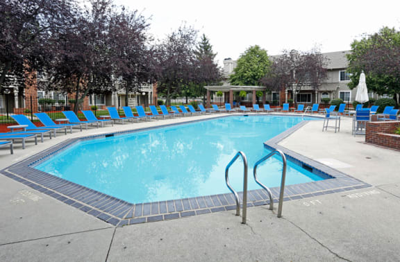 Swimming pool with ladder, cement pool deck with blue lounge chairs surrounding.  Buildings in background.