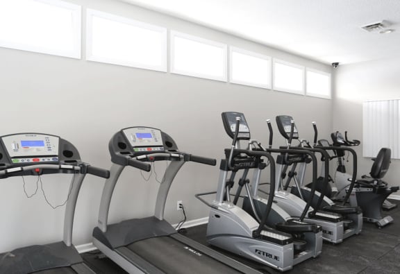 A row of 2 treadmills, and 2 elupticals along with another machine in the corner.  Windows across top by ceiling, white walls.