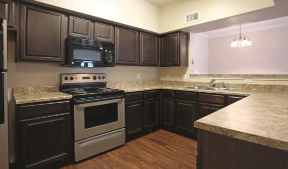 Dark wood cabinetry, stainless oven with black microwave.  Wood flooring and beige counter-top.  Light over sink.