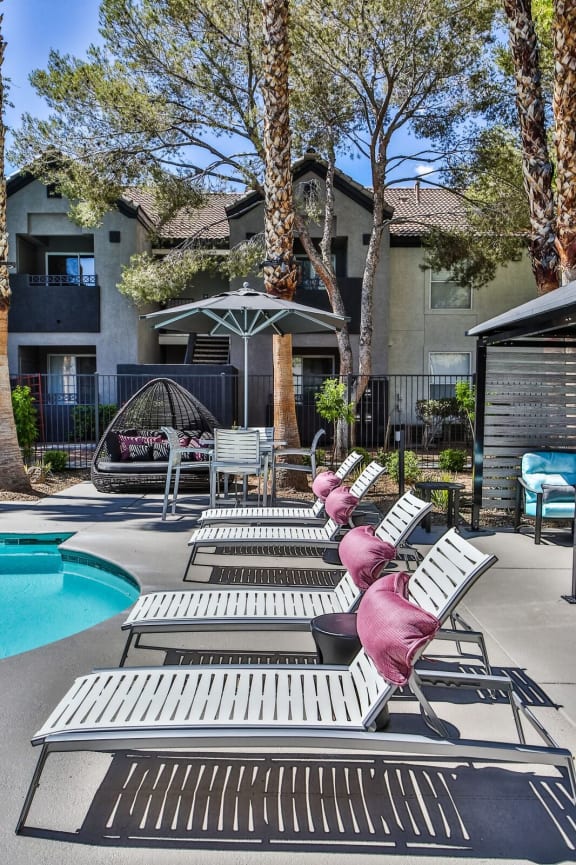Pool deck with lounge chairs with pillows and apartments in the background.