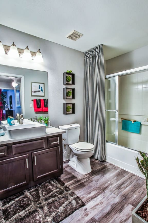 Bathroom with dark cabinets, light counter top, white sink, wood like style flooring, white toilet, and a white shower/tub combo.