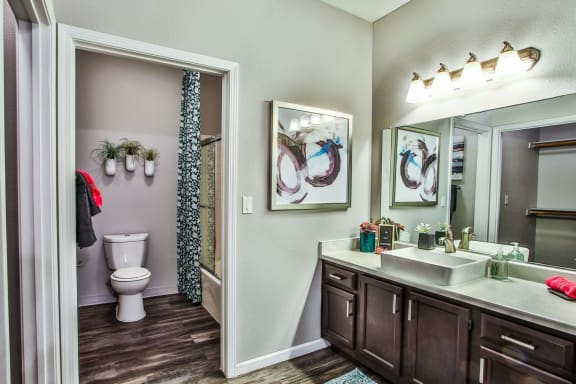 Bathroom with dark counters, light counter top, white sink, separate toilet/shower area, silver fixtures and wood like style flooring.
