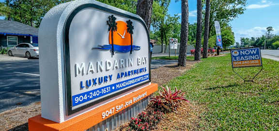 Street sign at 9047 San Jose Blvd. Jacksonville, FL 32257 that reads "Mandarin Bay Luxury Apartments" next to row of tall trees and "now leasing" sign.