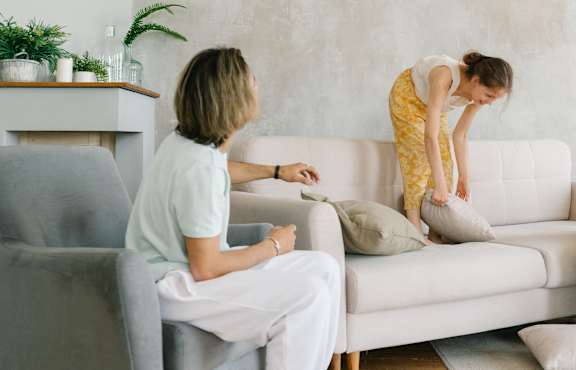 An elderly couple is sitting on a grey couch, with the woman holding a smartphone.