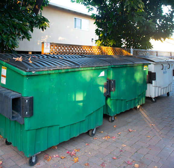 A green dumpster is parked on a brick sidewalk.