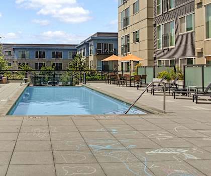 The turquoise outdoor pool with pool chairs, tables, and chairs with the apartments in the background at Leilani Apartment Homes. 
