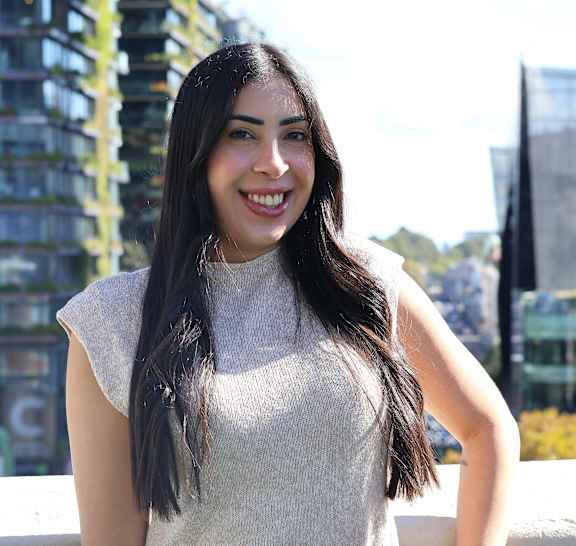 A woman with long hair is smiling and wearing a white top and a brown belt.