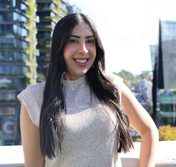 A woman with long hair is smiling and wearing a white top and a brown belt.