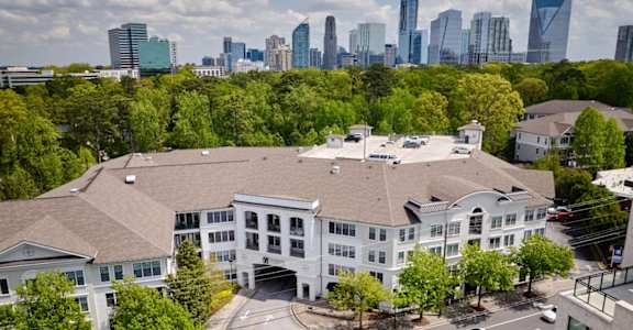 A large white building with a brown roof is surrounded by trees and has a city skyline in the background.