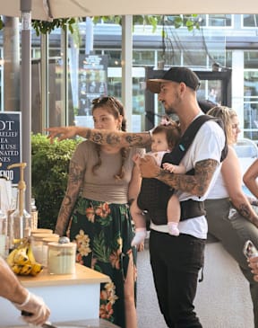 A woman with tattoos is holding a baby while standing next to a man with tattoos and a hat.