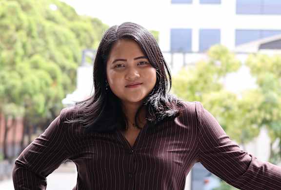 A woman with a black blazer and a gold necklace is standing in front of a red wall.