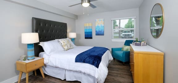 Mandarin bay apartment bedroom in Jacksonville, FL with large bed, sitting chair, window with slat blinds, and midcentury wooden dresser.