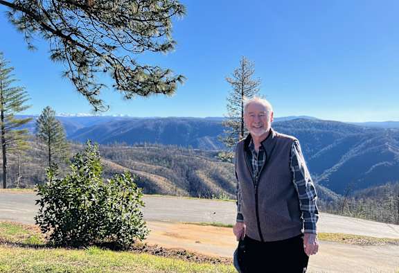 A man in a grey vest and plaid shirt stands in front of a scenic view of mountains.