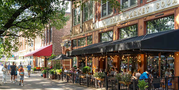 A street view of a market square with people walking and sitting at tables.