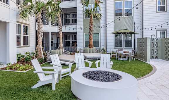 A white fire pit sits in the middle of a grassy area with white chairs around it.