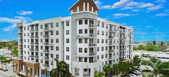 A large white building with a brown roof and balconies is surrounded by palm trees.