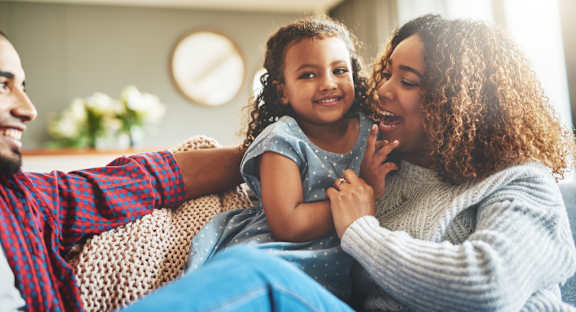 A family of three is sitting on a couch, with a clock on the wall behind them.