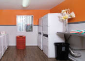 Thumbnail 21 of 23 - On site laundry at Fifteen 50 apartments in Las Vegas, 89119 with four washers and dryers, red trash can, and modern orange, blue, and white walls.
