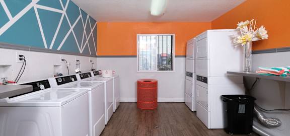 On site laundry at Fifteen 50 apartments in Las Vegas, 89119 with four washers and dryers, red trash can, and modern orange, blue, and white walls.