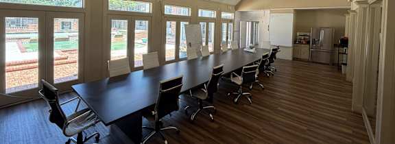 A conference room with a long table and chairs, large windows, and a view of a garden outside.