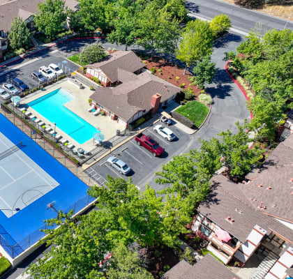 an aerial view of full size tennis court, swimming pool area and parking lot and apartment buildings surrounding. 