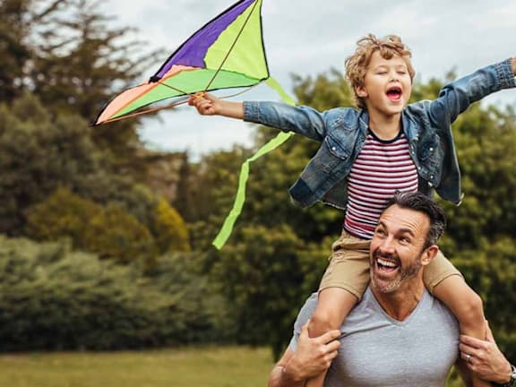 a man carrying a child on his shoulders while flying a kite
