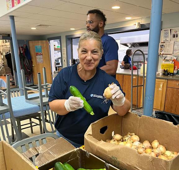 A woman in a blue shirt is holding a cucumber and a potato in a kitchen setting.