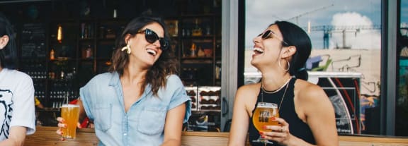 two women sitting on a bench laughing and drinking beer at Dey & Bergen, Harrison, 07029