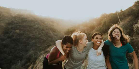 Four women are walking together on a path with a hill in the background.