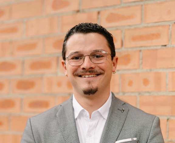 A man in a grey suit stands in front of a brick wall.