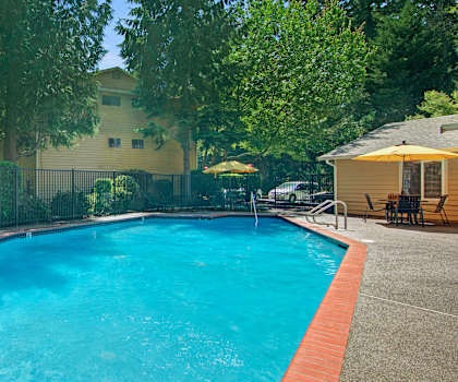 The large, turquoise outdoor pool with pool chairs, tables, and chairs with the apartments and the parking lot in the background at the Ridge and Shores Apartment Homes. 