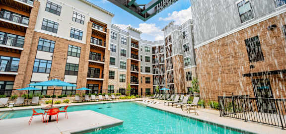 A swimming pool is surrounded by chairs and umbrellas in front of apartment buildings.