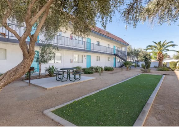 A white building with blue trim and a balcony is surrounded by a tree, a bench, and a green lawn.