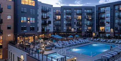 A large swimming pool surrounded by lounge chairs and umbrellas in front of apartment buildings.