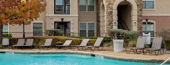 A swimming pool is surrounded by a concrete patio and a building with a stone pillar.