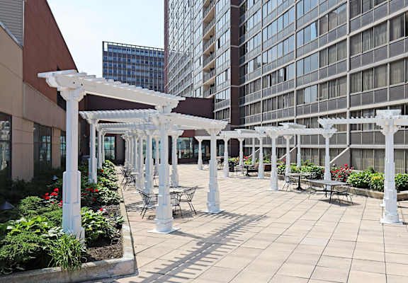 A patio with white pergolas and chairs is surrounded by buildings.