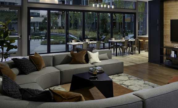 The amazing, modern resident lounge with a fireplace, a gray couch shaped in a U, decorations, large windows with curtains that show the outdoor courtyard, plush chairs, and coffee tables at Uptown at Kirkland Urban Apartment Homes. 