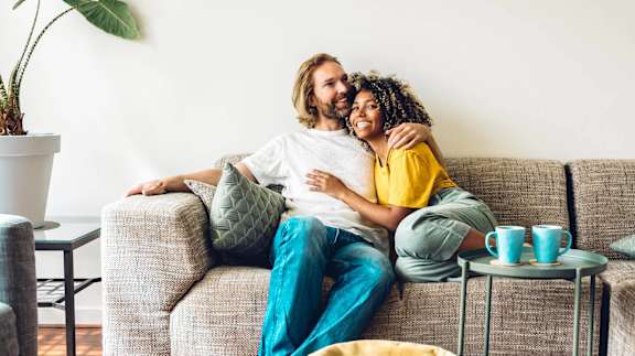 A couple is sitting on a couch with a potted plant to their left and a small table with cups on it to their right.