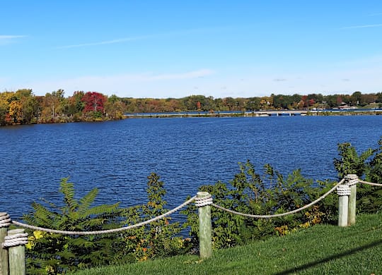 A lake surrounded by trees with a rope fence in the foreground at The Waverly on the Lake - Belleville, MI Apartment , Belleville, MI