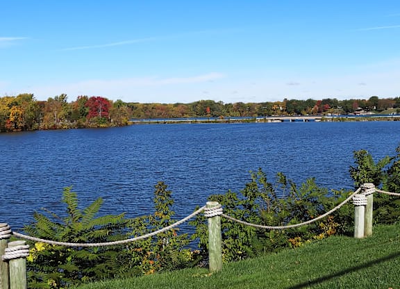 A lake surrounded by trees with a rope fence in the foreground at The Waverly on the Lake - Belleville, MI Apartment , Belleville, MI