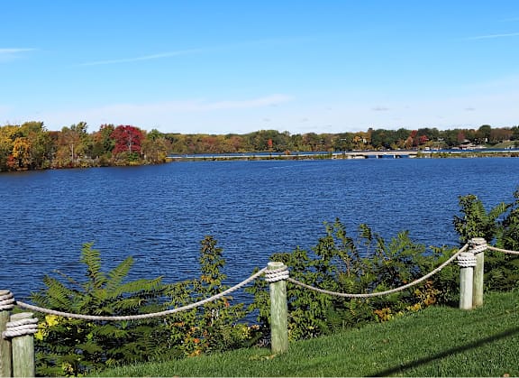 A lake surrounded by trees with a rope fence in the foreground at The Waverly on the Lake - Belleville, MI Apartment , Belleville, MI