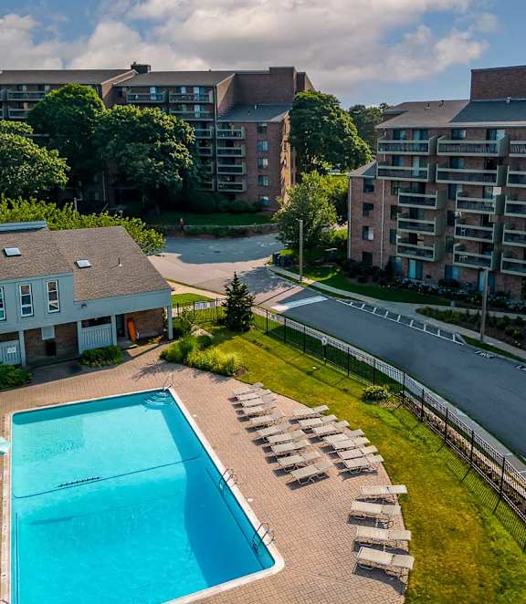 A swimming pool surrounded by a brick patio and a grassy area with a road and apartment buildings in the background.