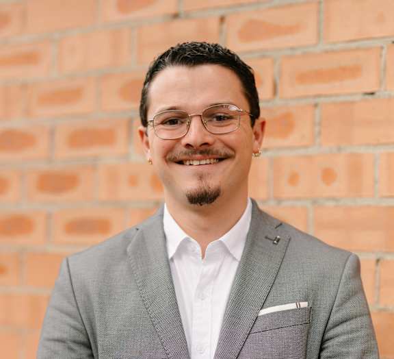 A man in a grey suit stands in front of a brick wall.