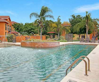 The large, turquoise outdoor pool with palm trees, a small fountain, pool chairs, tables, and chairs with the orange apartments in the background at the Retreat at Speedway Apartment Homes. 