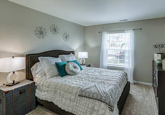 A bedroom with a bed, nightstands, and a window with a view of the city at Abbey Ridge Apartment Homes, Oxford