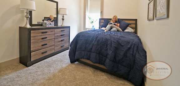 A woman is sitting on a bed with a blue comforter in a bedroom with a large mirror and a dresser with a lamp.