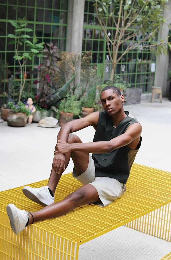 A man sits on a yellow mat in a greenhouse.