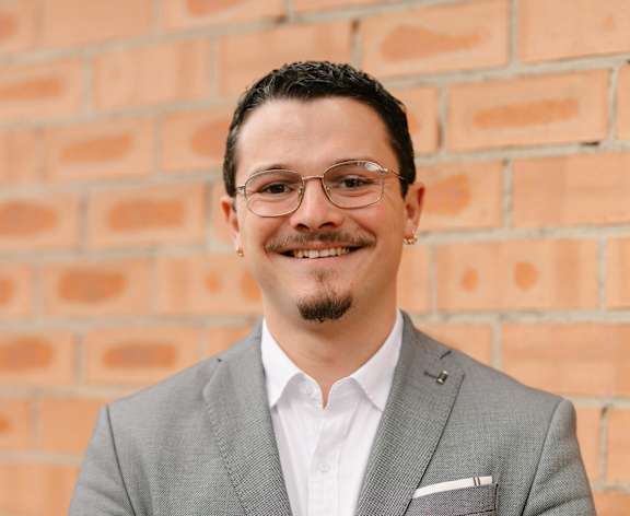 A man in a grey suit stands in front of a brick wall.